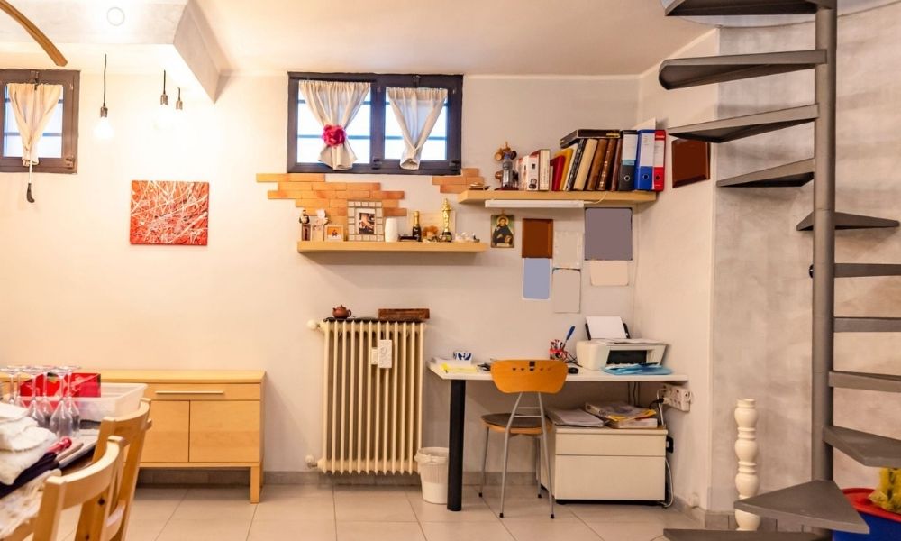 Sala con scala a chiocciola, tavolo ingombro e oggetti personali — lo spazio dello studio fotografico prima dell'intervento, privo di identità di brand