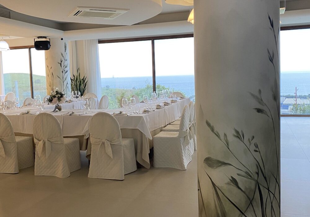 Pilastro di una location matrimoni rivestito con carta da parati botanica a motivo di erbe e uccelli, con profilo metallico alla base, sala allestita con tavolata imbandita e vista sul mare che continua oltre il pilastro