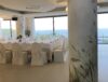 Pilastro di una location matrimoni rivestito con carta da parati botanica a motivo di erbe e uccelli, con profilo metallico alla base, sala allestita con tavolata imbandita e vista sul mare che continua oltre il pilastro