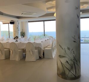 Pilastro di una location matrimoni rivestito con carta da parati botanica a motivo di erbe e uccelli, con profilo metallico alla base, sala allestita con tavolata imbandita e vista sul mare che continua oltre il pilastro