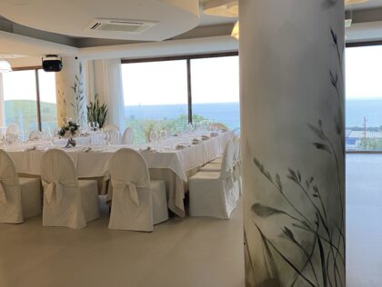 Pilastro di una location matrimoni rivestito con carta da parati botanica a motivo di erbe e uccelli, con profilo metallico alla base, sala allestita con tavolata imbandita e vista sul mare che continua oltre il pilastro